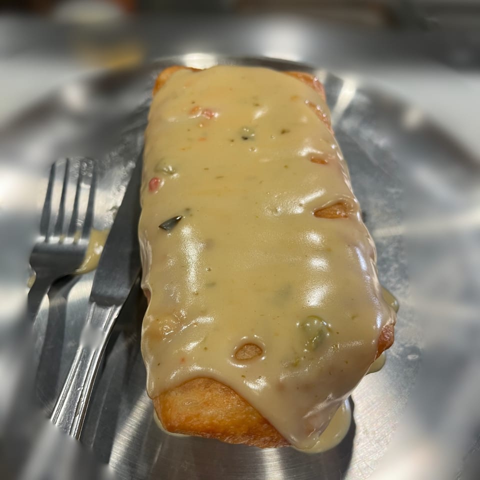A golden-brown chimichanga covered in creamy white sauce with a fork and knife on a metal tray.