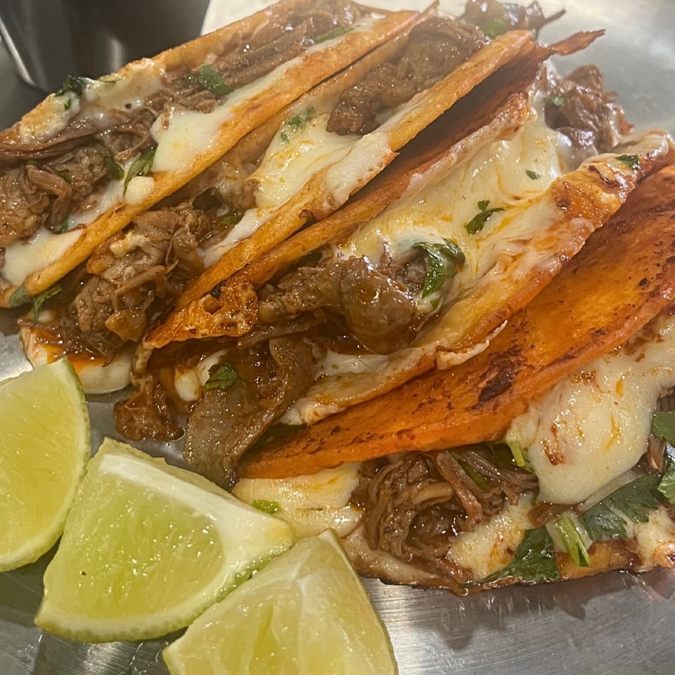 Close-up of three cheesy birria tacos with shredded beef and lime wedges on a metal plate.
