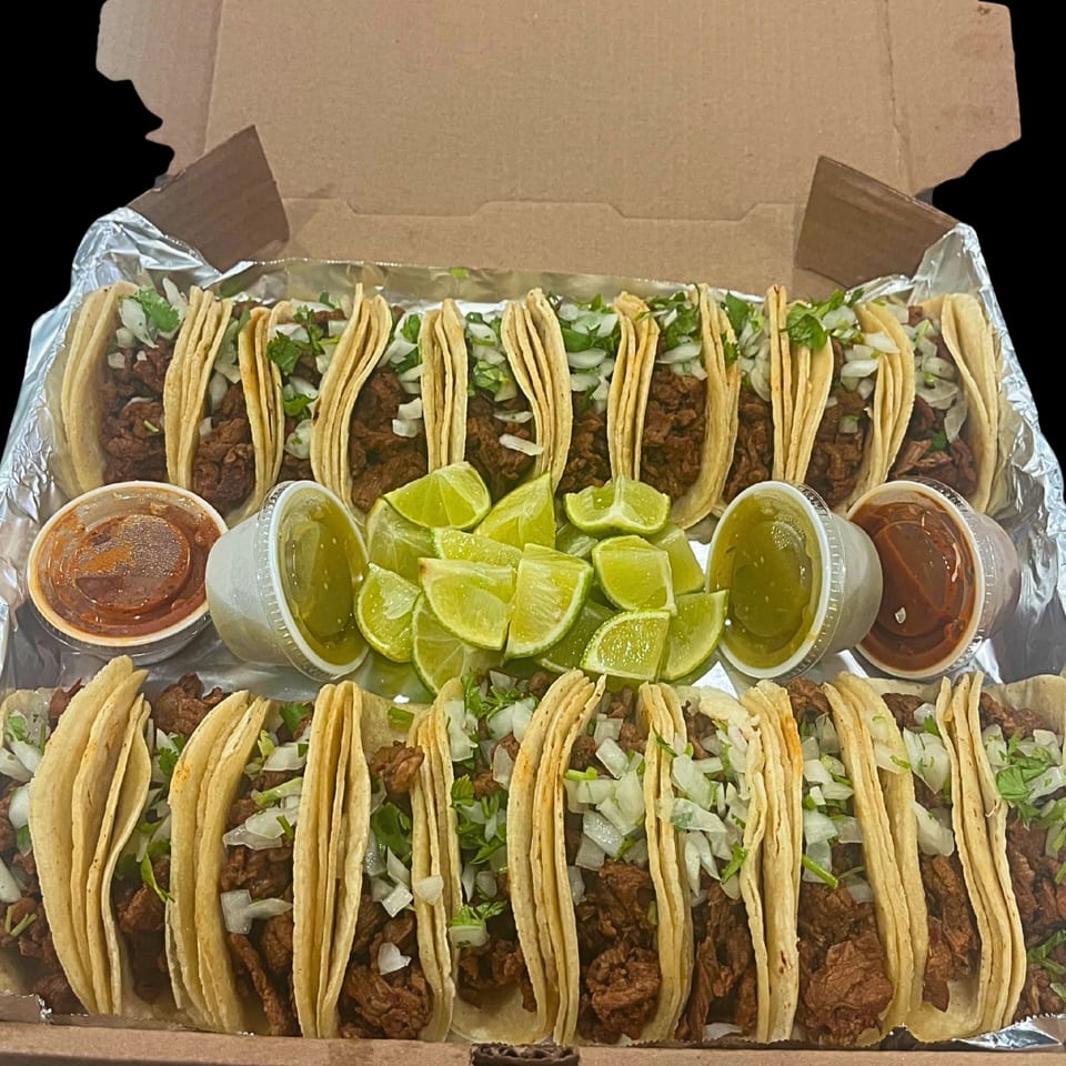 A dozen Family Grill Mexican street tacos with meat, onions, cilantro, lime wedges, and two sauces in a box.