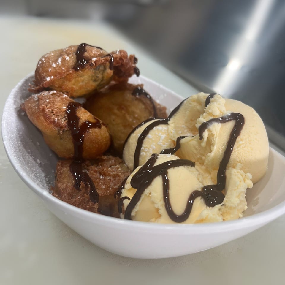 Deep-fried Mexican dessert with powdered sugar, chocolate drizzle, and vanilla ice cream in a white bowl.