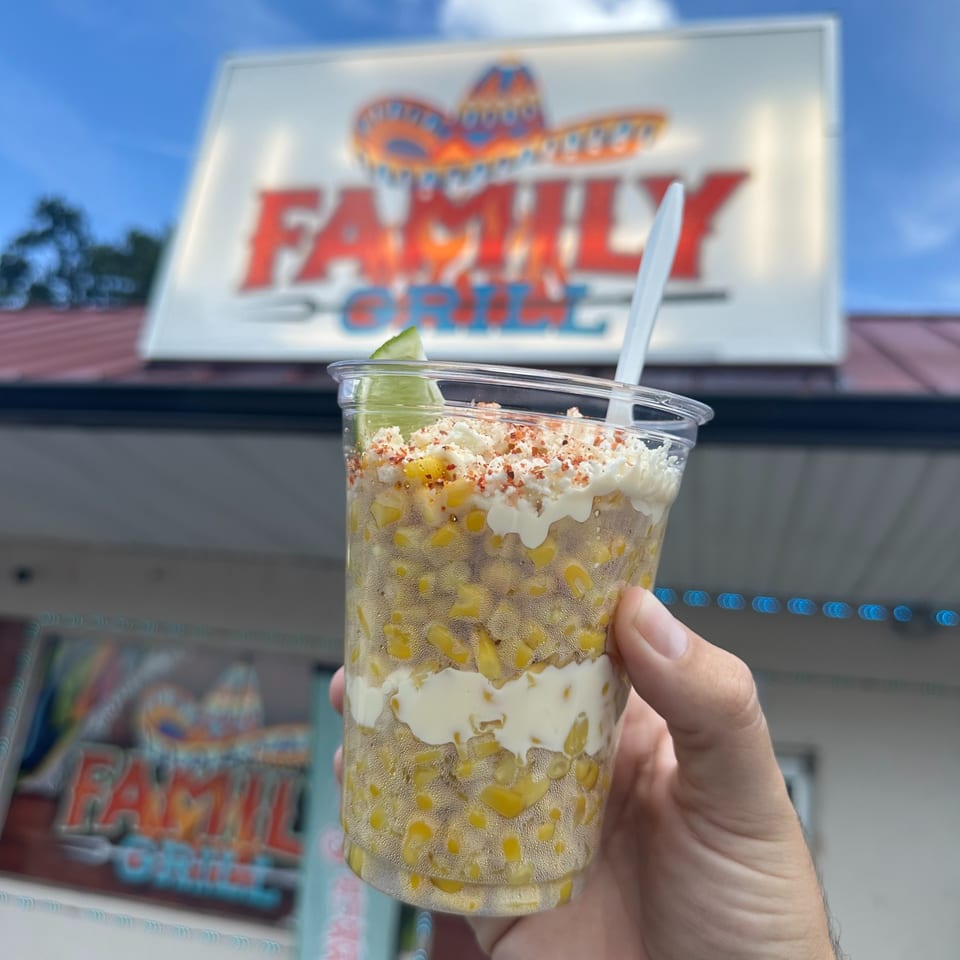 Hand holding a cup of Mexican street corn with lime, cheese, and chili powder in front of the Family Grill sign.